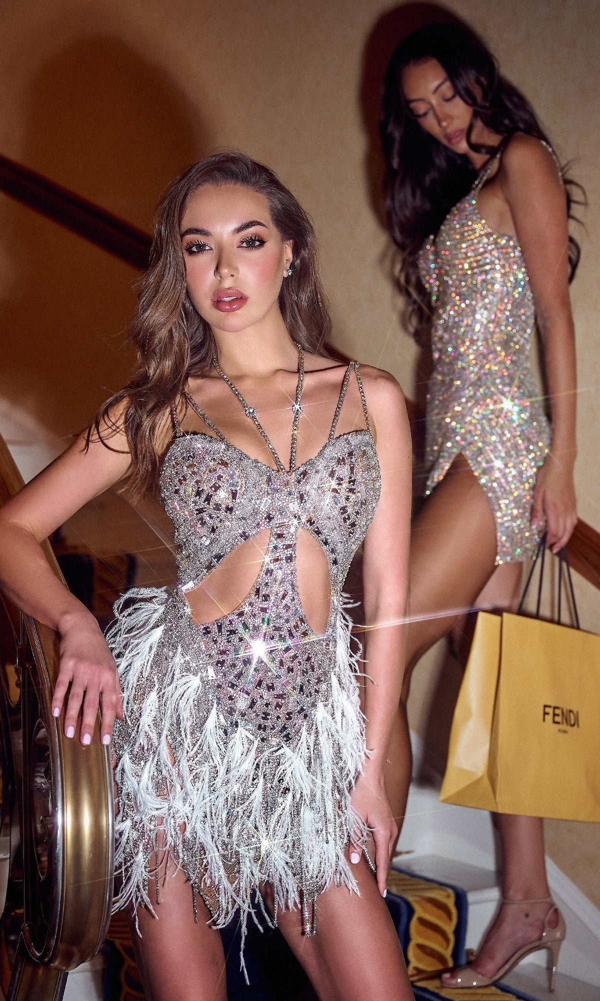 Two women in sparkly Portia and Scarlett dresses stand on a staircase; one poses in a beaded fringe dress with feathered details, the Short Homecoming Dress: Portia and Scarlett PS24898C, while the other holds a Fendi shopping bag.